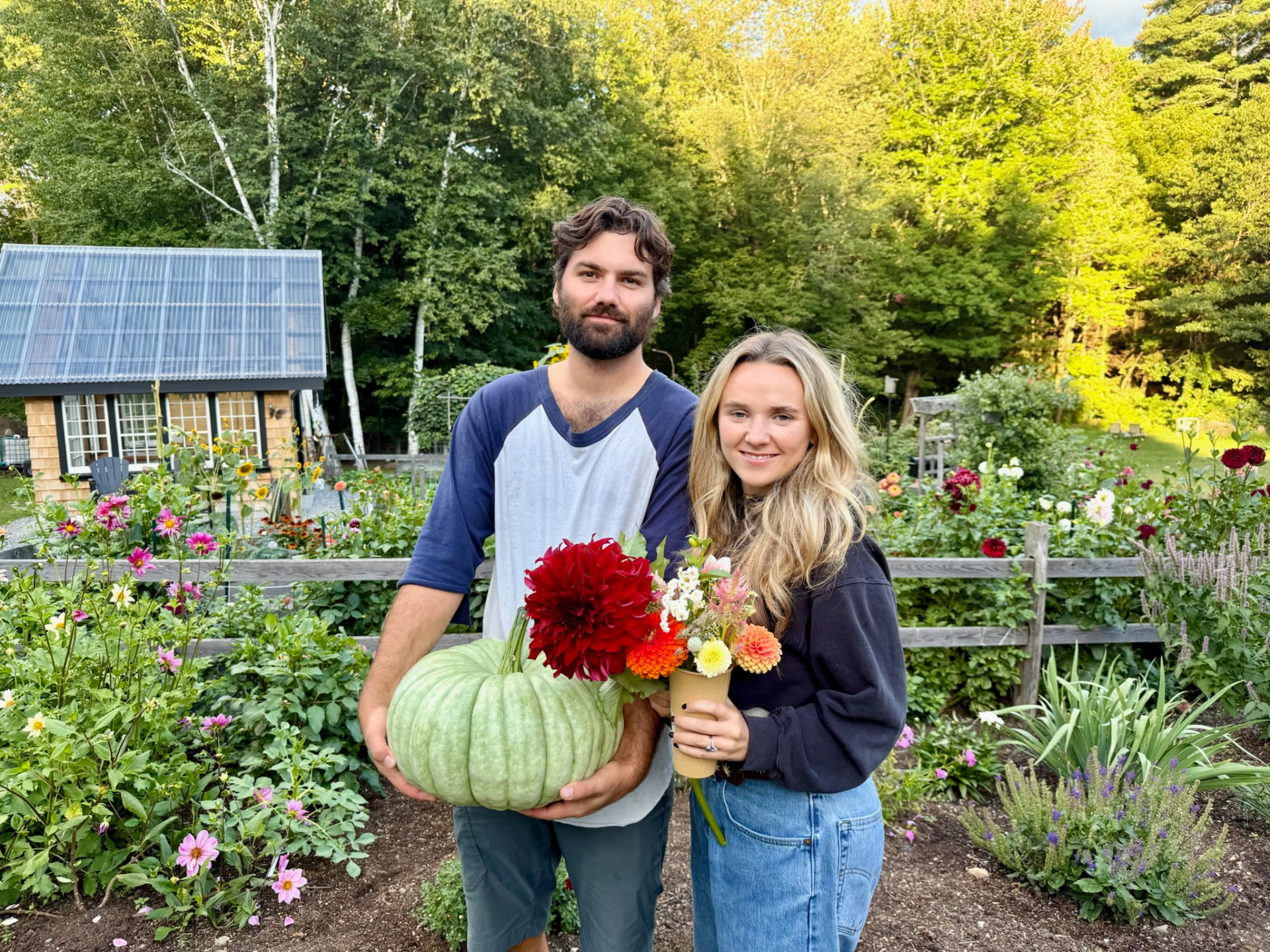 A couple posing in the flower rows holding a homegrown pumpkin and a wrapped bouquet